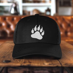 May include: Black trucker hat with a white bear paw print on the front. The hat has a curved brim and a mesh back. The paw print is centered on the front panel.