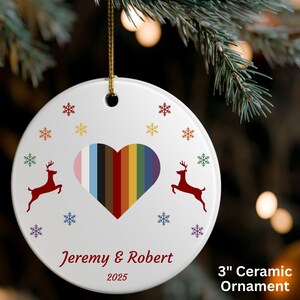 May include: A white ceramic Christmas ornament featuring a heart-shaped design with rainbow stripes. Two red reindeer and snowflakes decorate the ornament. The names "Jeremy & Robert" and the year "2025" are printed below. The ornament is 7.6 cm in diameter.