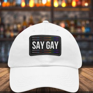 Op de afbeelding: Witte baseballpet met een zwart embleem met de tekst "SAY GAY" in witte letters. Het embleem heeft horizontale lijnen in regenboogkleuren. De pet staat op een houten oppervlak met een vage achtergrond.