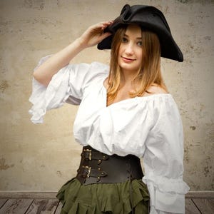 Könnte beinhalten: Eine Frau trägt eine weiße Bauernbluse mit einem braunen Ledergürtel und einem schwarzen Dreispitz. Sie trägt einen grünen Rock mit Rüschen.