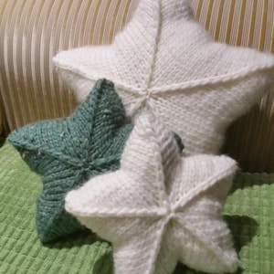 May include: Three star-shaped pillows with a chunky knit texture. One large white pillow is positioned at the back, while a smaller white pillow and a medium-sized sage green pillow with subtle flecks of color are arranged in the front.