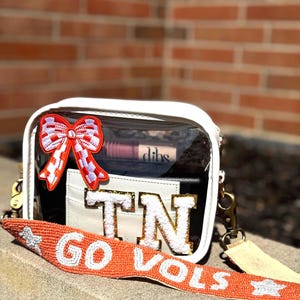 May include: A clear, white-trimmed crossbody bag with a red and white bow patch. The bag features a beaded orange strap with "GO VOLS" text and a "TN" patch. The bag contains a wallet and a lipstick.
