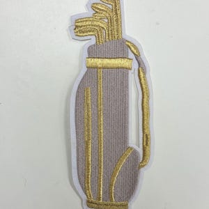 May include: A gray golf bag with gold embroidered details. The bag is filled with gold embroidered golf clubs.