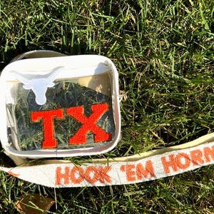 May include: A clear, rectangular crossbody bag with a white strap and orange text that reads "HOOK 'EM HORNS." The bag features a white longhorn logo and the letters "TX" in orange. The bag is on a bed of green grass.