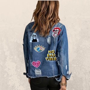 May include: A medium-wash denim jacket with distressed details and patches. Patches include a woman in sunglasses, an eye with a wave, a heart, and a red tongue. The jacket has the words "NO BAD VIBES" in yellow.