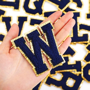 May include: A navy blue felt letter W with a gold glitter outline. The letter is held in a hand.