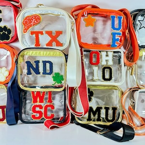 May include: A collection of clear plastic crossbody bags with various college team logos and patches. The bags are lined up in a row and feature logos for teams such as UC, AU, ND, Wisconsin, Ohio State, Elon, and Tennessee.
