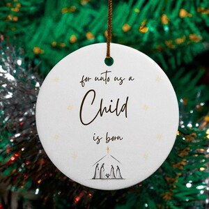May include: White ceramic Christmas ornament with the text "for unto us a Child is born" in elegant script. The ornament features a small illustration of a nativity scene and is suspended by a brown string.