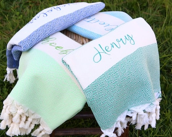 Può includere: Tre asciugamani personalizzati con frange. Gli asciugamani sono piegati e impilati su uno sgabello di legno. Gli asciugamani sono verdi, blu e bianchi con motivi a rombi. I nomi "Henry", "Chloe" e "Joy" sono ricamati sugli asciugamani.