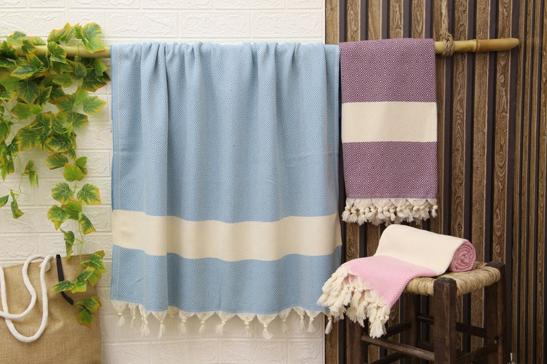 Pu&ograve; includere: Tre asciugamani turchi, uno blu chiaro con una striscia bianca, uno rosa con una striscia bianca e uno viola con una striscia bianca. Gli asciugamani sono appesi a un'asta di legno e sono in cotone.