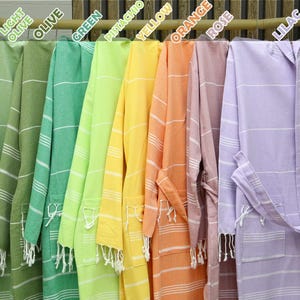 May include: A collection of ten Turkish towels, each in a different color, hanging on a wooden rack. The colors are mint, light olive, olive, green, pistachio, yellow, orange, rose, and lilac.