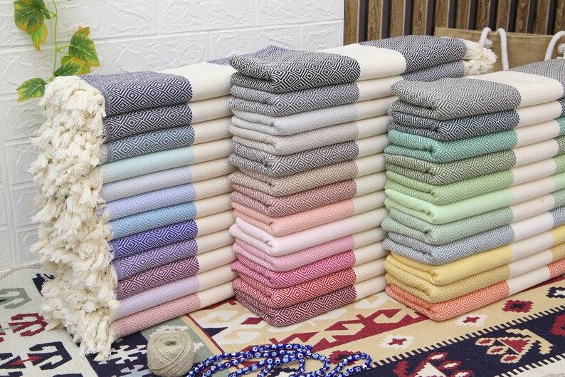 Pu&ograve; includere: Una pila di asciugamani turchi colorati con un motivo a rombi. Gli asciugamani sono piegati e impilati in una variet&agrave; di colori, tra cui blu, verde, rosa e giallo.