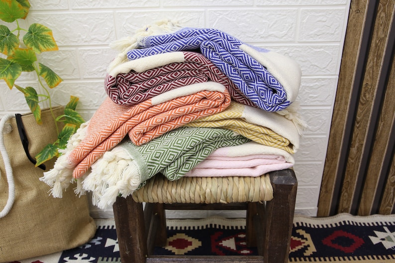 Pu&ograve; includere: Una pila di sei asciugamani turchi colorati con un motivo a rombi, piegati e disposti su uno sgabello di legno. Gli asciugamani sono in tonalit&agrave; di blu, rosso, arancio, verde e rosa.