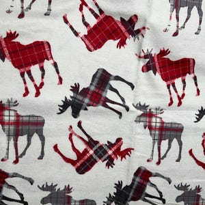 May include: White fabric with a repeating pattern of moose silhouettes in red, black, and gray plaid. The moose are in various poses, creating a rustic and festive design. Ideal for home decor or seasonal crafts.
