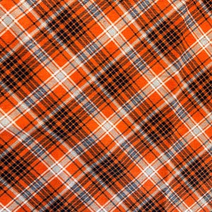 May include: Close-up of a fabric with a plaid pattern. The design features a repeating pattern of orange, black, white, and gray intersecting lines. The fabric has a woven texture, creating a visually appealing and textured surface.