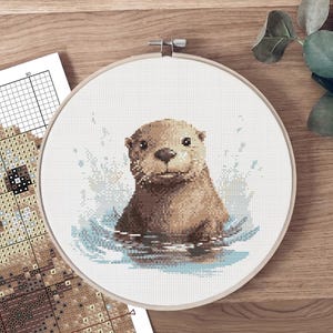 Pode incluir: Bordado em ponto cruz de uma lontra num aro redondo. A lontra é castanha com a face clara, emergindo da água azul. Um gráfico de padrões é visível. O bordado está sobre uma superfície de madeira.