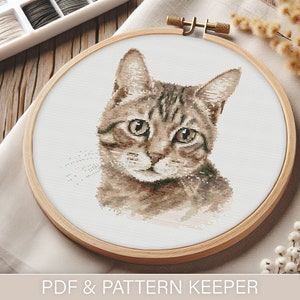 Op de afbeelding: Een kruissteekpatroon van een bruine tabbykat met groene ogen, ingelijst in een houten borduurring. De ring ligt op een beige stoffen oppervlak.