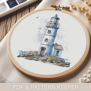 May include: A white and blue lighthouse cross-stitch pattern on a white fabric stretched over a wooden embroidery hoop. The lighthouse is on a rocky shore with a small building attached to it.