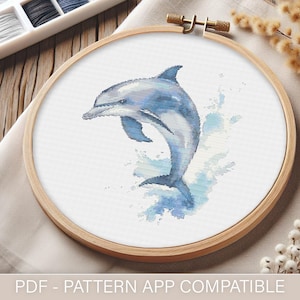 May include: A cross-stitch pattern featuring a watercolour-style illustration of a dolphin jumping out of the water. The pattern is in a wooden embroidery hoop and is ready to be stitched.