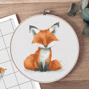Può includere: Ricamo a punto croce di una volpe seduta con gli occhi chiusi, nei toni dell'arancione, bianco e grigio, in un telaio di legno. Un foglio modello con una griglia e un disegno di volpe è visibile a sinistra. Lo sfondo è una superficie di legno.