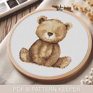 Könnte beinhalten: Ein Kreuzstich-Muster eines braunen Teddybären in einem hölzernen Stickrahmen. Der Bär sitzt mit geschlossenen Augen und einem kleinen Lächeln. Der Rahmen liegt auf einem weißen Tuch und einer Holzoberfläche.