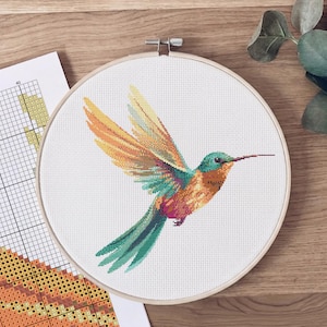 May include: A completed cross-stitch of a hummingbird in flight, featuring orange, yellow, green, and purple hues. The embroidery is set in a wooden hoop, with a pattern chart visible nearby. The craft project is on a wooden surface.