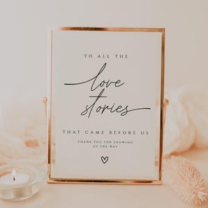 May include: A framed sign with gold edging, displaying the text "To All The Love Stories That Came Before Us" in a stylish script. A small heart symbol is at the bottom. A candle and decorative item are in front.