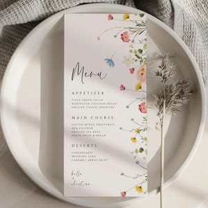 May include: A wedding menu card with a floral design. The menu features a handwritten "Menu" title and a list of appetizers, main courses, and desserts. The card is decorated with watercolor flowers in pink, yellow, and blue. The names "Belle + Christian" are at the bottom.