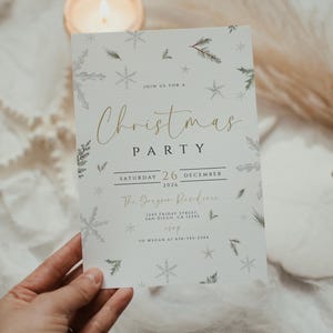 Op de afbeelding: Een kerstfeestuitnodiging met gouden scripttekst en illustraties van sneeuwvlokken en dennentakken. De uitnodiging bevat de datum, tijd en locatiegegevens. De uitnodiging wordt door een hand vastgehouden.