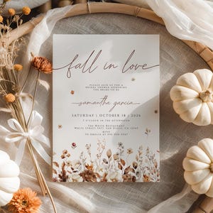 以下が含まれることがあります： 「fall in love」という言葉がエレガントな筆記体で書かれたブライダルシャワーの招待状。茶色、ベージュ、白の色合いの花柄と植物のイラストが特徴です。詳細には、花嫁の名前、日付、時間、場所が含まれています。