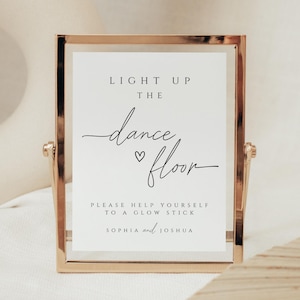 Può includere: Un cartello con cornice dorata con la scritta "LIGHT UP THE dance floor" in corsivo nero. Sotto, si legge "PLEASE HELP YOURSELF TO A GLOW STICK" e i nomi "SOPHIA and JOSHUA". Il cartello è su sfondo bianco.