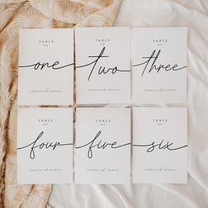 Könnte beinhalten: Sechs weiße Tischkarten mit schwarzen Schriftnummern. Jede Karte zeigt eine Zahl von eins bis sechs, mit den Worten "Table No." über der Zahl. Die Namen "Sophia and Joshua" sind darunter gedruckt. Die Karten sind auf einem neutralen Stoff angeordnet.