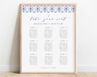 May include: A framed seating chart for Madeline's Baptism, featuring a decorative blue and white tile design at the top. The words "take your seat" are written in elegant script above the name and event details. The chart lists guest names under table numbers.
