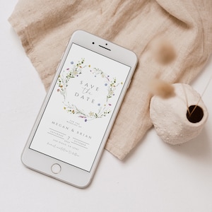 Puede incluir: Un teléfono inteligente blanco que muestra una tarjeta de guardar la fecha con un diseño de corona floral. El texto dice "Save the Date" para la boda de Megan & Brian el sábado 7 de octubre de 2034 en San Diego, CA 12349.