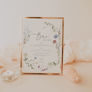 May include: A framed sign with a floral border and the text "The Bar Menu". The menu lists beer, wine, and signature cocktails. The sign is on a white surface with a candle and a decorative piece.