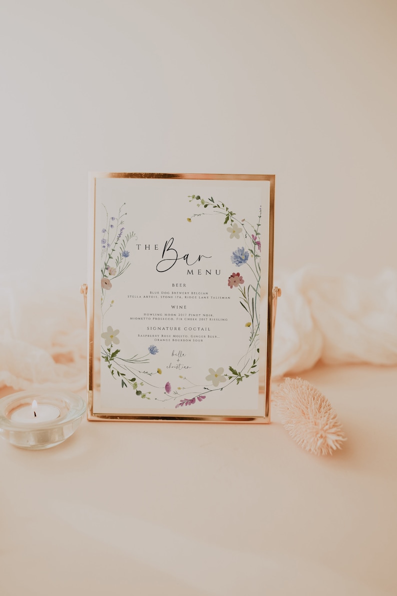 May include: A framed sign with a floral border and the text "The Bar Menu". The menu lists beer, wine, and signature cocktails. The sign is on a white surface with a candle and a decorative piece.