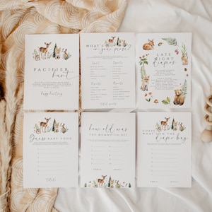 May include: A collection of six baby shower game cards with woodland animal illustrations. The cards feature games like "Pacifier Hunt," "What's in Your Purse?" and "Late Night Diapers." A wooden rattle is also visible.