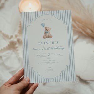 May include: A baby's first birthday invitation card with a blue and white striped design. The card features a teddy bear illustration and the text "OLIVER'S beary first birthday". The card includes the date, time, and location details.