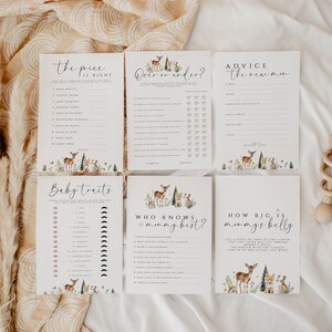 May include: A collection of baby shower game cards with woodland animal illustrations. The cards include games like "The Price is Right," "Baby Traits," and "How Big is Mommy's Belly?" A wooden baby rattle is also visible.