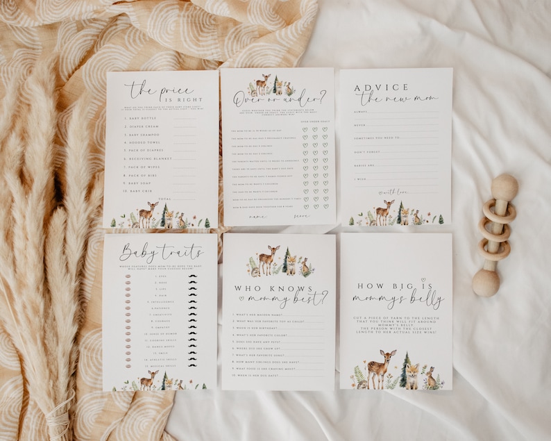 May include: A collection of baby shower game cards with woodland animal illustrations. The cards include games like "The Price is Right," "Baby Traits," and "How Big is Mommy's Belly?" A wooden baby rattle is also visible.
