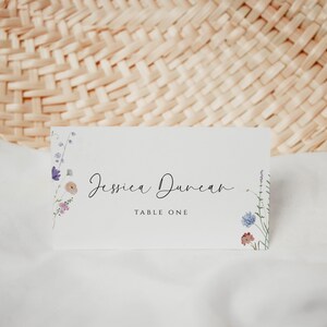May include: A white place card with a floral design on the left and right sides. The text "Jessica Duncan" is written in black cursive script. The text "Table One" is written in black below the name.