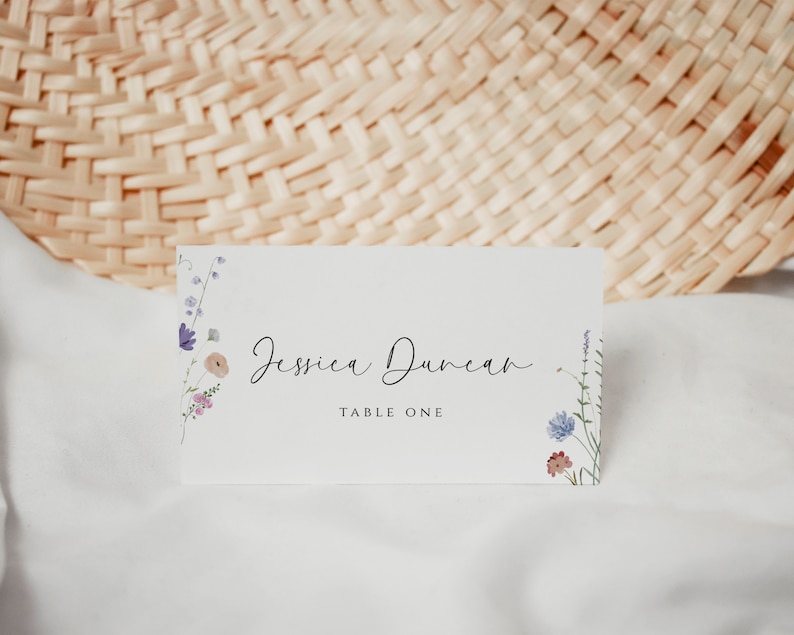 May include: A white place card with a floral design on the left and right sides. The text "Jessica Duncan" is written in black cursive script. The text "Table One" is written in black below the name.