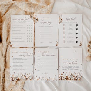 May include: A set of six baby shower game cards with floral and leaf accents. The cards have titles like "Over or Under?" and "Guess What's in the Diaper Bag?" A wooden rattle is also present.