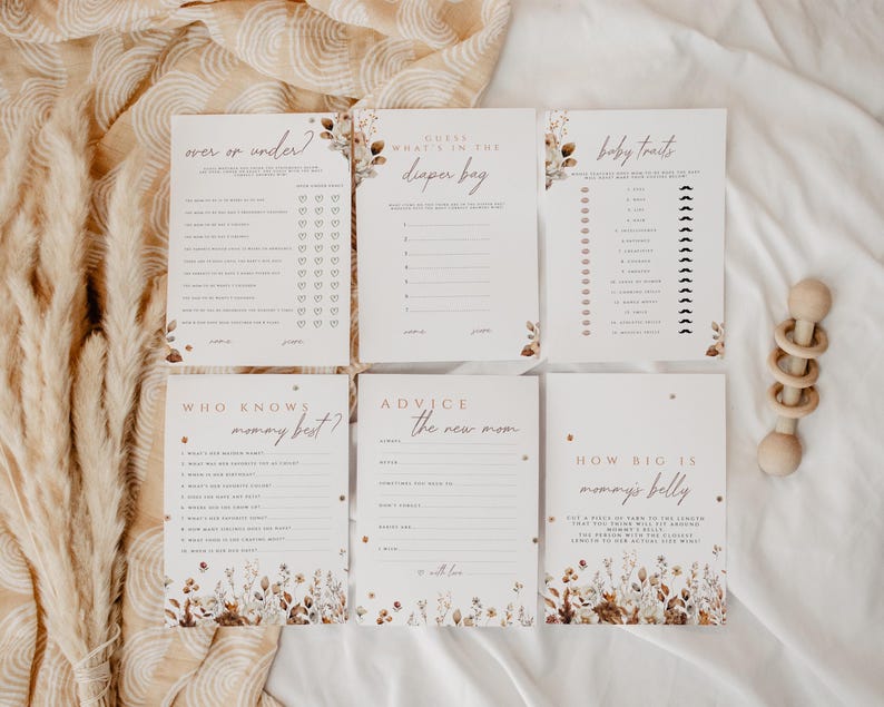 May include: A set of six baby shower game cards with floral and leaf accents. The cards have titles like "Over or Under?" and "Guess What's in the Diaper Bag?" A wooden rattle is also present.