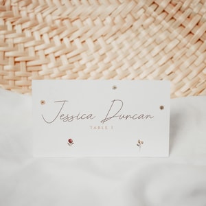 May include: A white place card with the name "Jessica Duncan" in a cursive font, with the text "TABLE 1" below. The card is decorated with small floral accents and rests on a white surface, with a woven background.