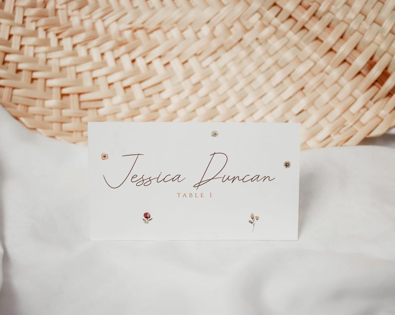 May include: A white place card with the name "Jessica Duncan" in a cursive font, with the text "TABLE 1" below. The card is decorated with small floral accents and rests on a white surface, with a woven background.