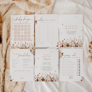 May include: A collection of baby shower game cards with floral accents and soft color palettes. The cards include games like "Baby Bingo," "Who's That Baby?" and "Diaper Raffle." A wooden rattle is also visible. The overall theme is a rustic, bohemian style.