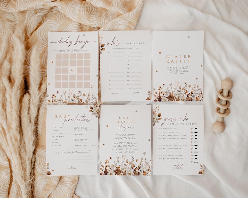 May include: A collection of baby shower game cards with floral accents and soft color palettes. The cards include games like "Baby Bingo," "Who's That Baby?" and "Diaper Raffle." A wooden rattle is also visible. The overall theme is a rustic, bohemian style.