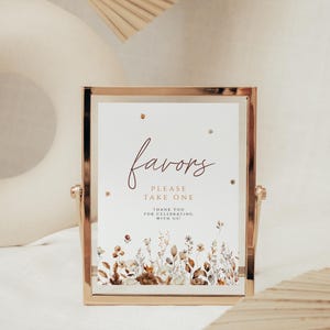 May include: A gold-framed sign with the word "favors" in elegant script. The sign reads "Please take one" and "Thank you for celebrating with us!" Floral illustrations in muted tones adorn the bottom edge, creating a rustic aesthetic.