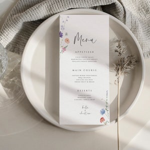 May include: A white menu card with a floral design and the text "Menu", "Appetizer", "Field Green Salad", "Marinated Chicken Breast", "Grilled Cheese Cro&ucirc;tons", "Main Course", "Roasted Mixed Vegetables", "Grilled Sea Bass", "Fresh Fruit & Bread", "Desserts", "Cheesecake", "Wedding Cake", "Caramel Apple Crisp", "Belle + Christian" printed on it.